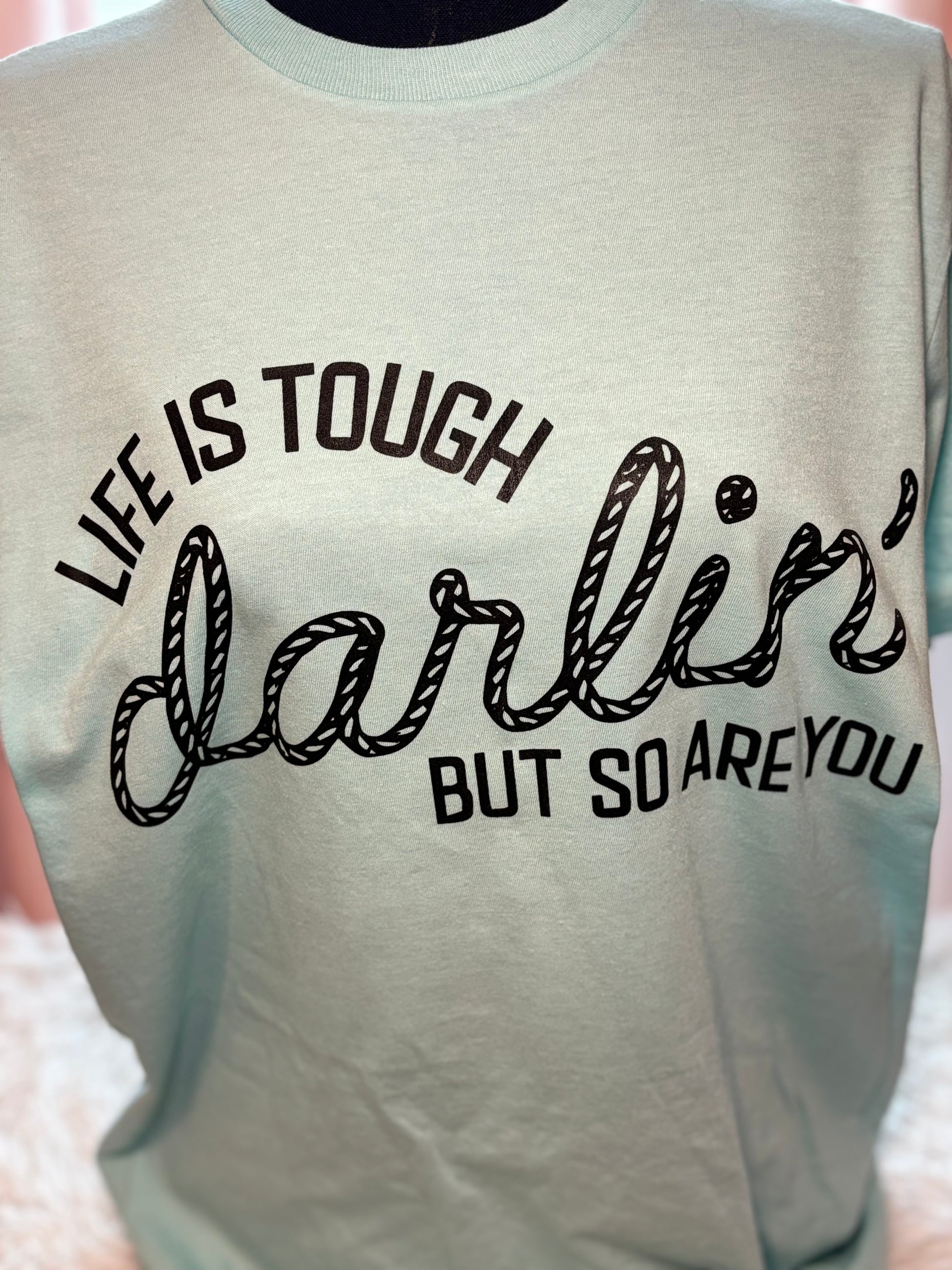 Light blue heat press t-shirt with motivational quote on a textured surface- Life is tough darlin but so are you