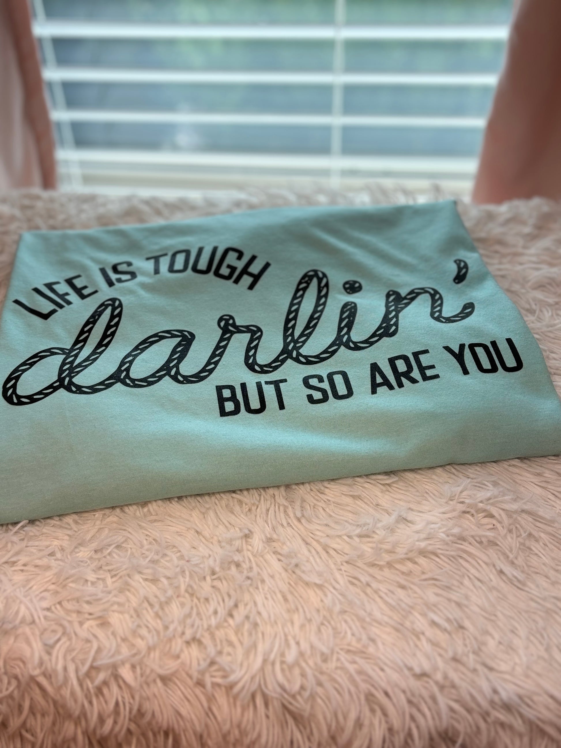 Light blue heat press t-shirt with motivational quote on a textured surface- Life is tough darlin but so are you