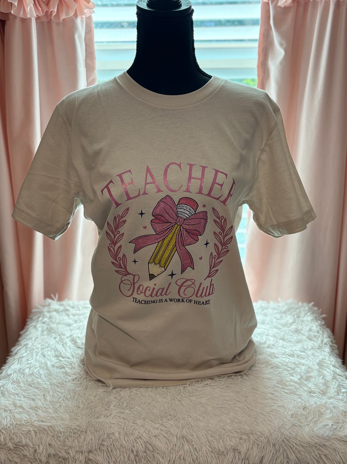 Beige heat press T-shirt with glitter 'Teacher Social Club' design featuring a pink bow and pencil on a light background.