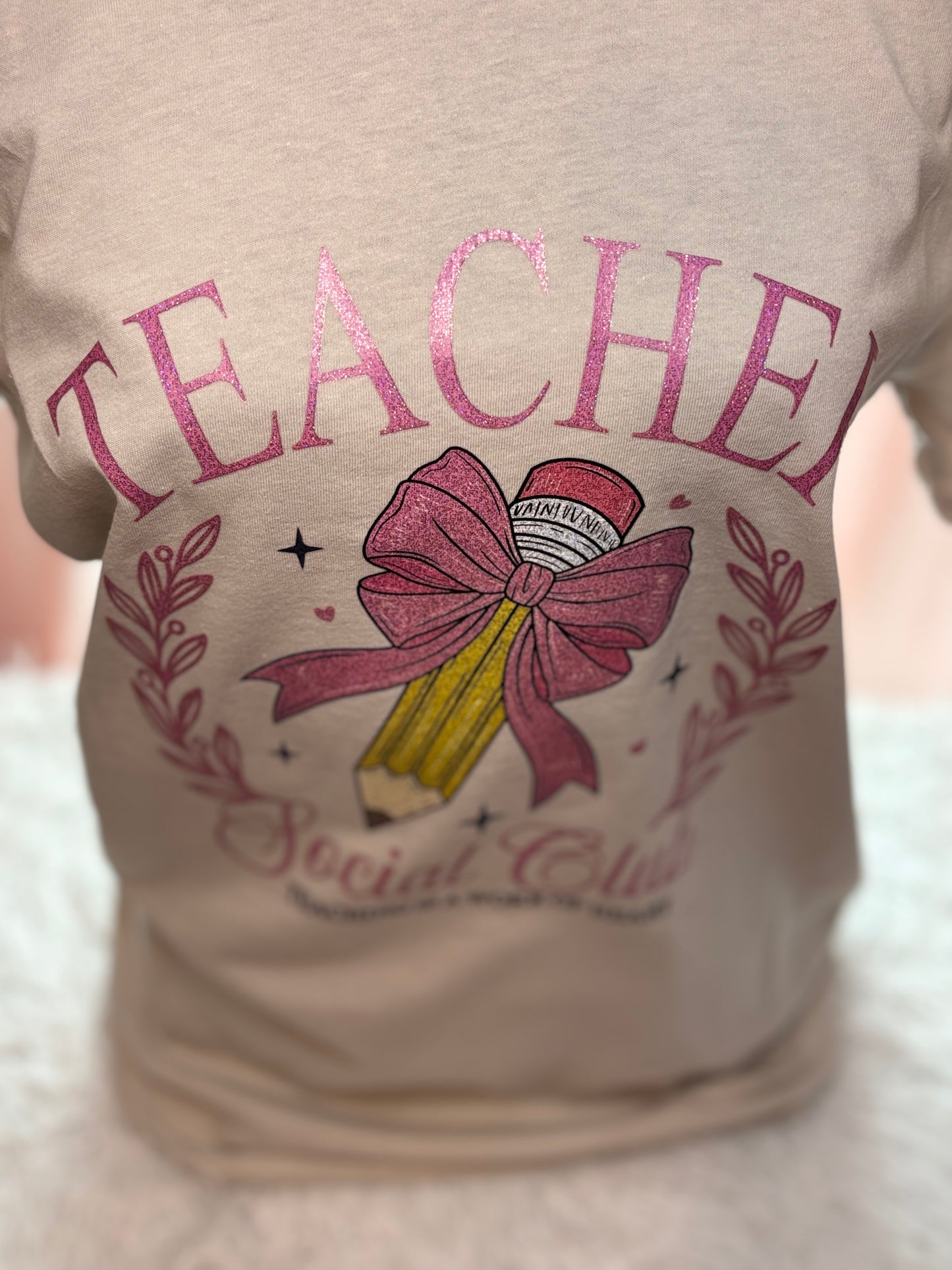 Beige heat press T-shirt with glitter 'Teacher Social Club' design featuring a pink bow and pencil on a light background.