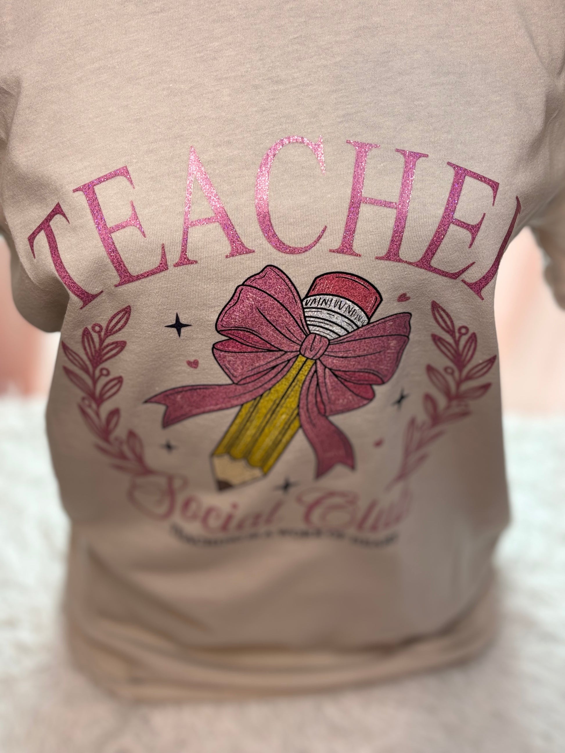 Beige heat press T-shirt with glitter 'Teacher Social Club' design featuring a pink bow and pencil on a light background.