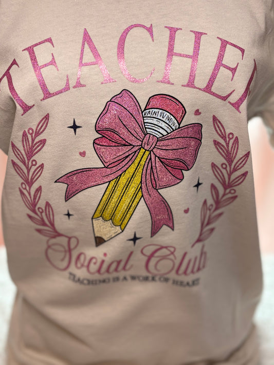 Beige heat press T-shirt with glitter 'Teacher Social Club' design featuring a pink bow and pencil on a light background.