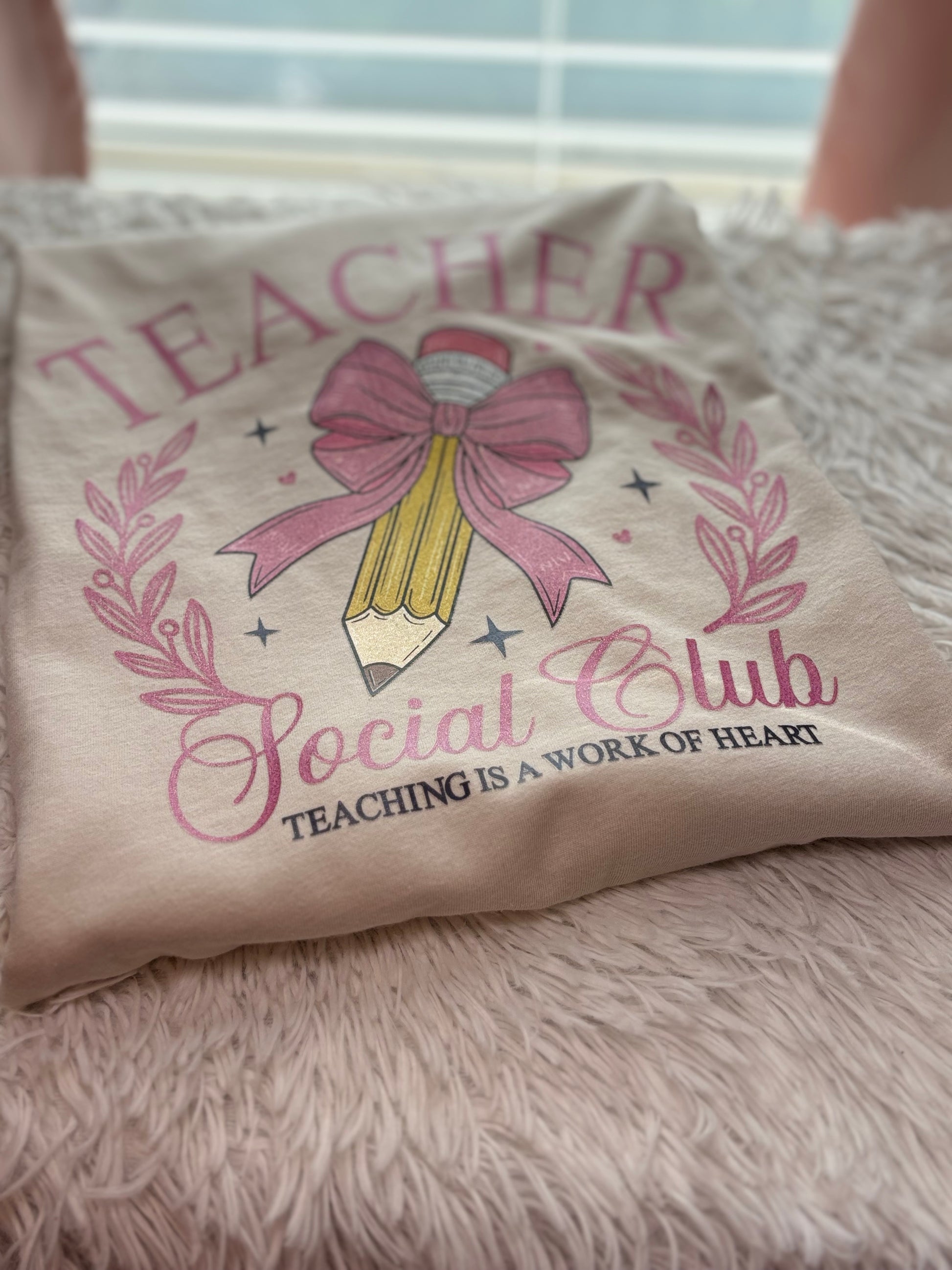 Beige heat press T-shirt with glitter 'Teacher Social Club' design featuring a pink bow and pencil on a light background.