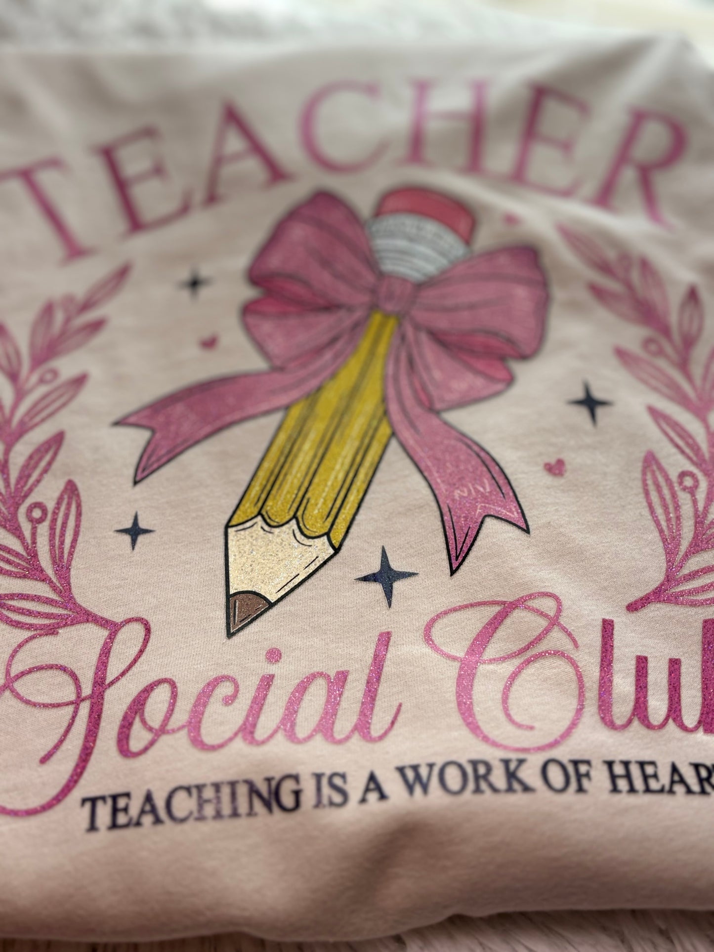 Beige heat press T-shirt with glitter 'Teacher Social Club' design featuring a pink bow and pencil on a light background.