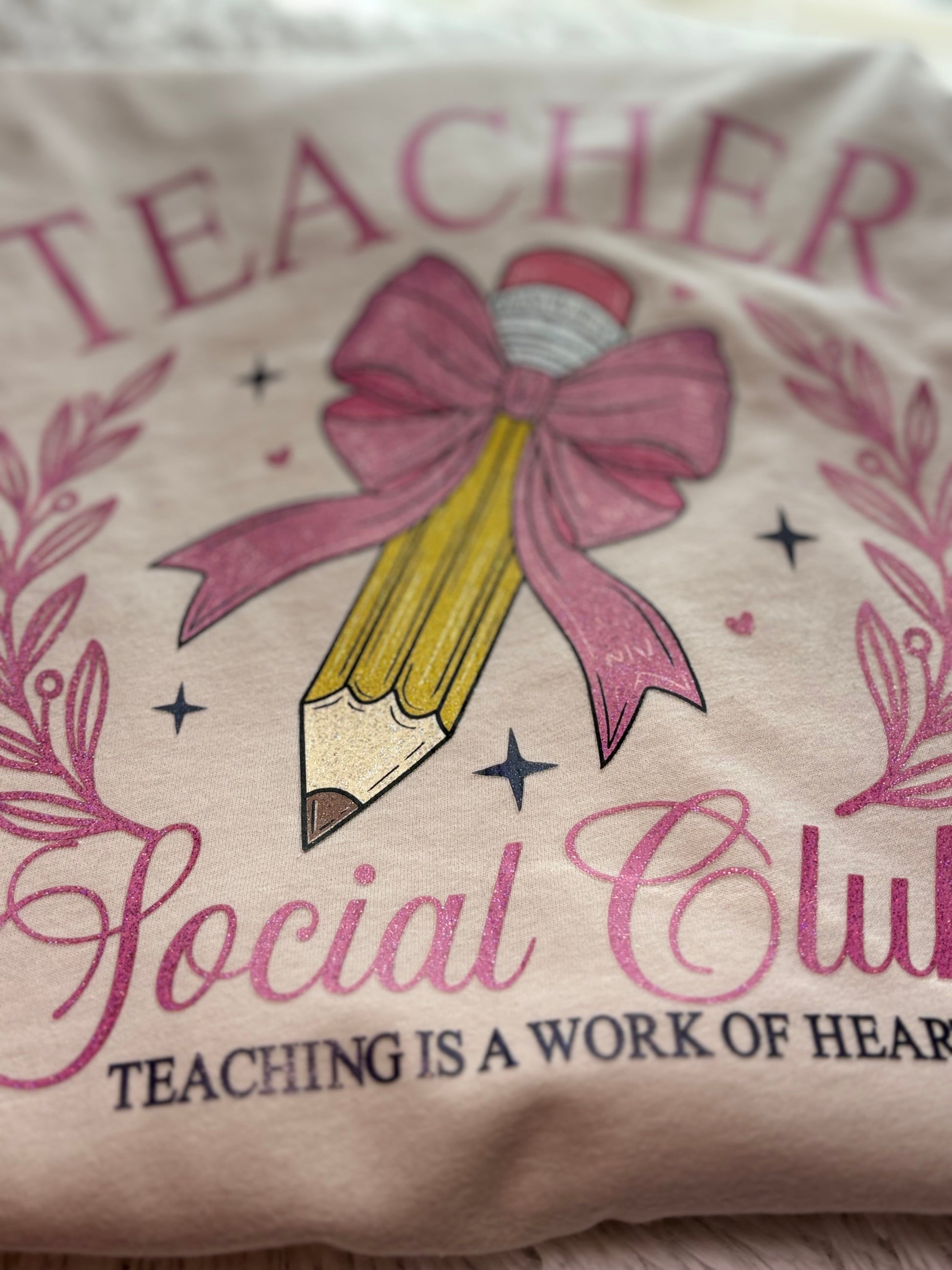 Beige heat press T-shirt with glitter 'Teacher Social Club' design featuring a pink bow and pencil on a light background.