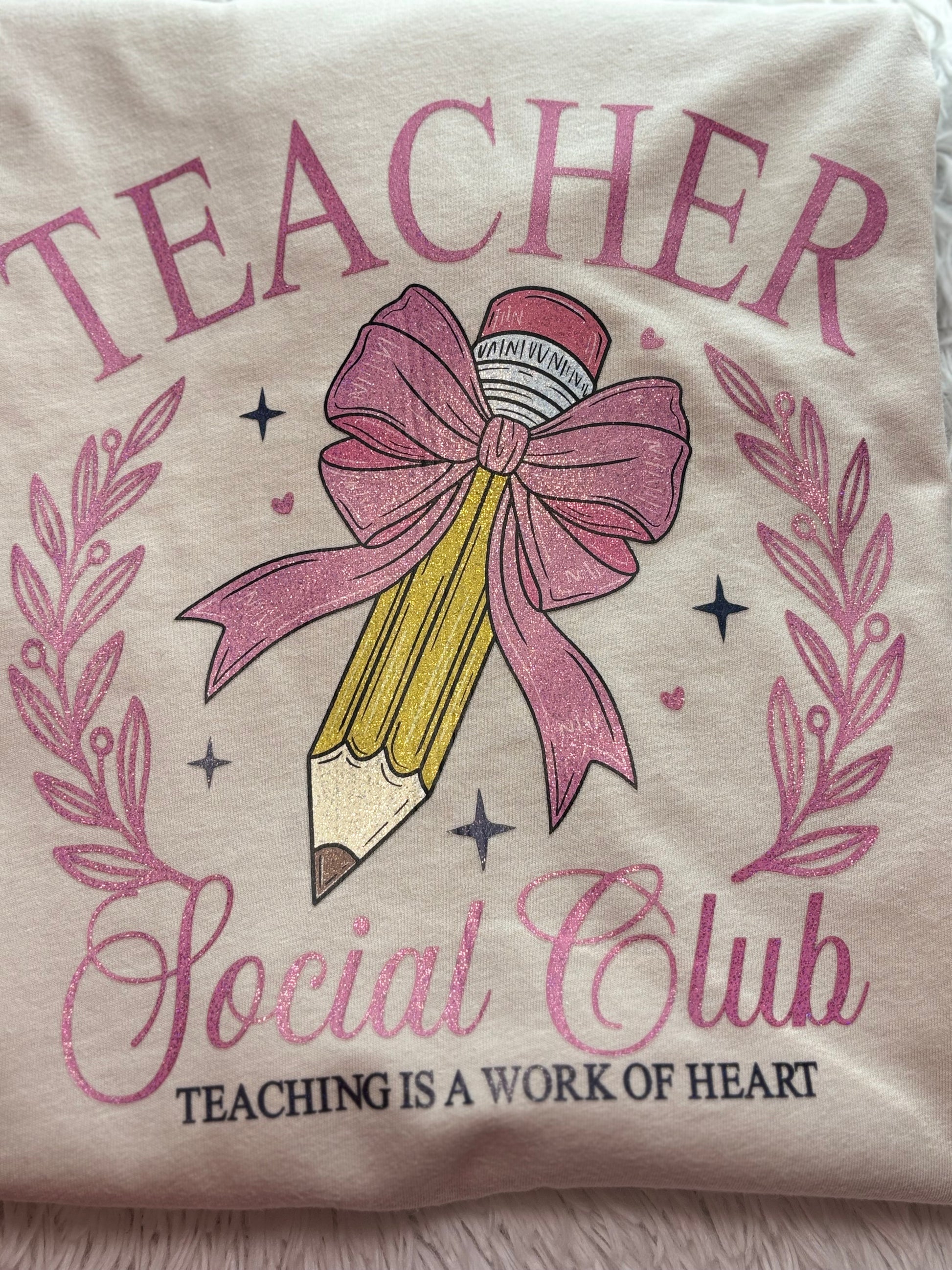 Beige heat press T-shirt with glitter 'Teacher Social Club' design featuring a pink bow and pencil on a light background.