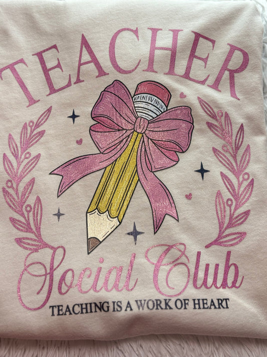 Beige heat press T-shirt with glitter 'Teacher Social Club' design featuring a pink bow and pencil on a light background.