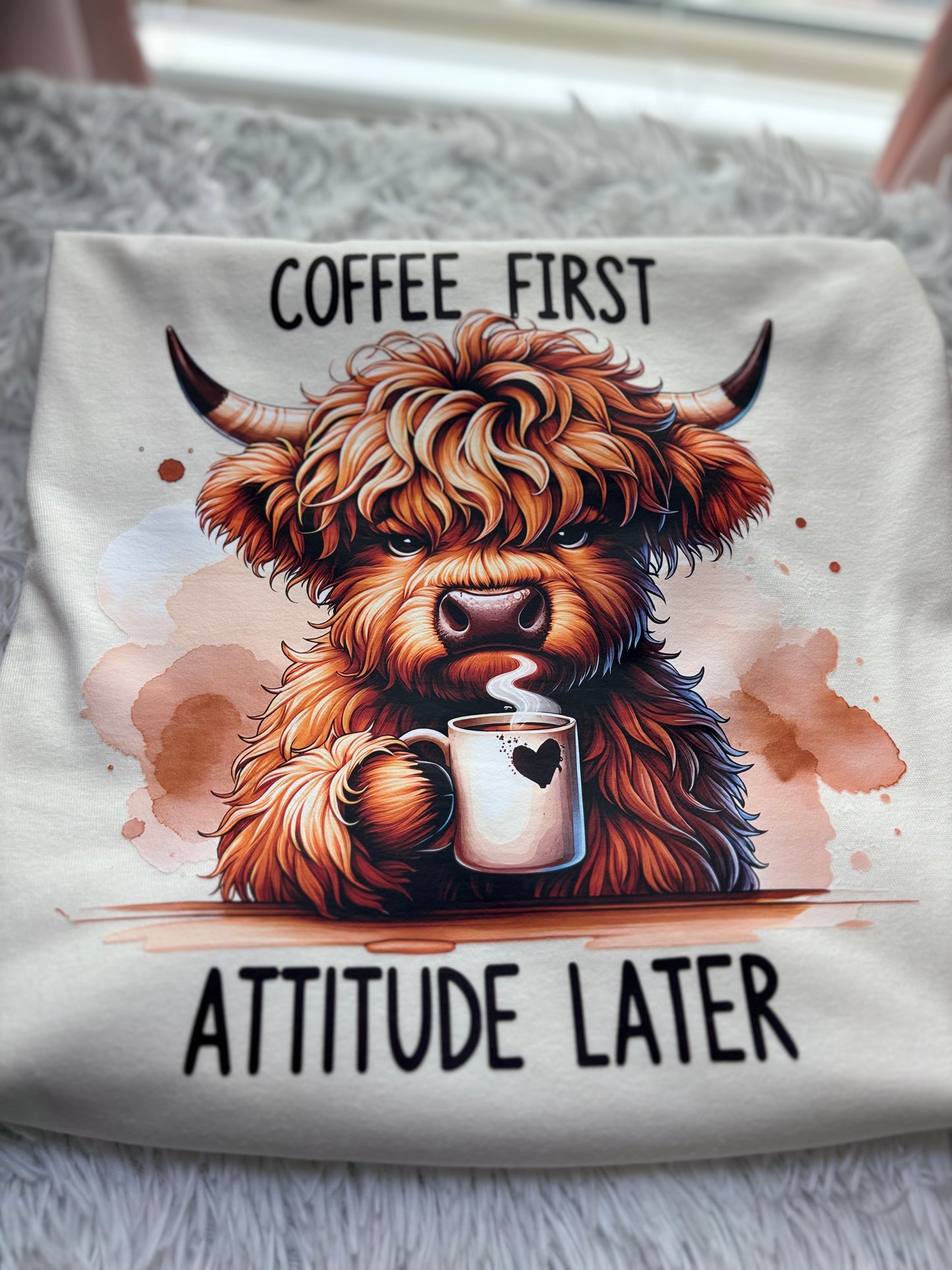 Beige heat press t-shirt with a Highland cow holding a coffee cup and text 'Coffee First, Attitude Later'