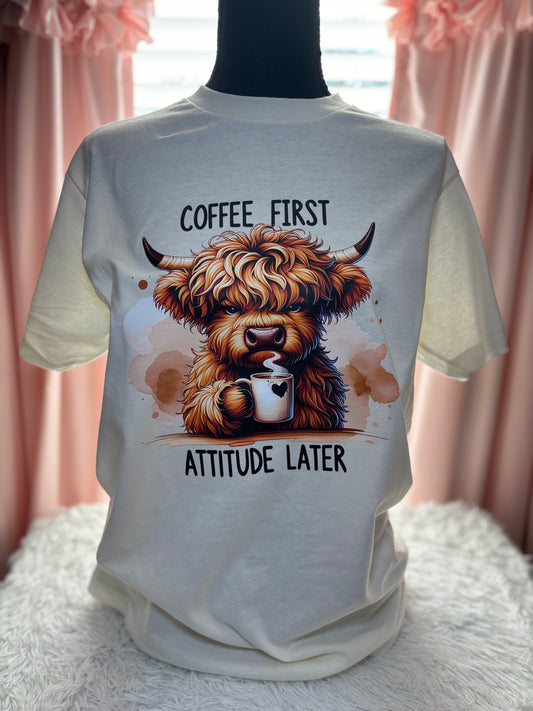 Beige heat press t-shirt with a Highland cow holding a coffee cup and text 'Coffee First, Attitude Later'