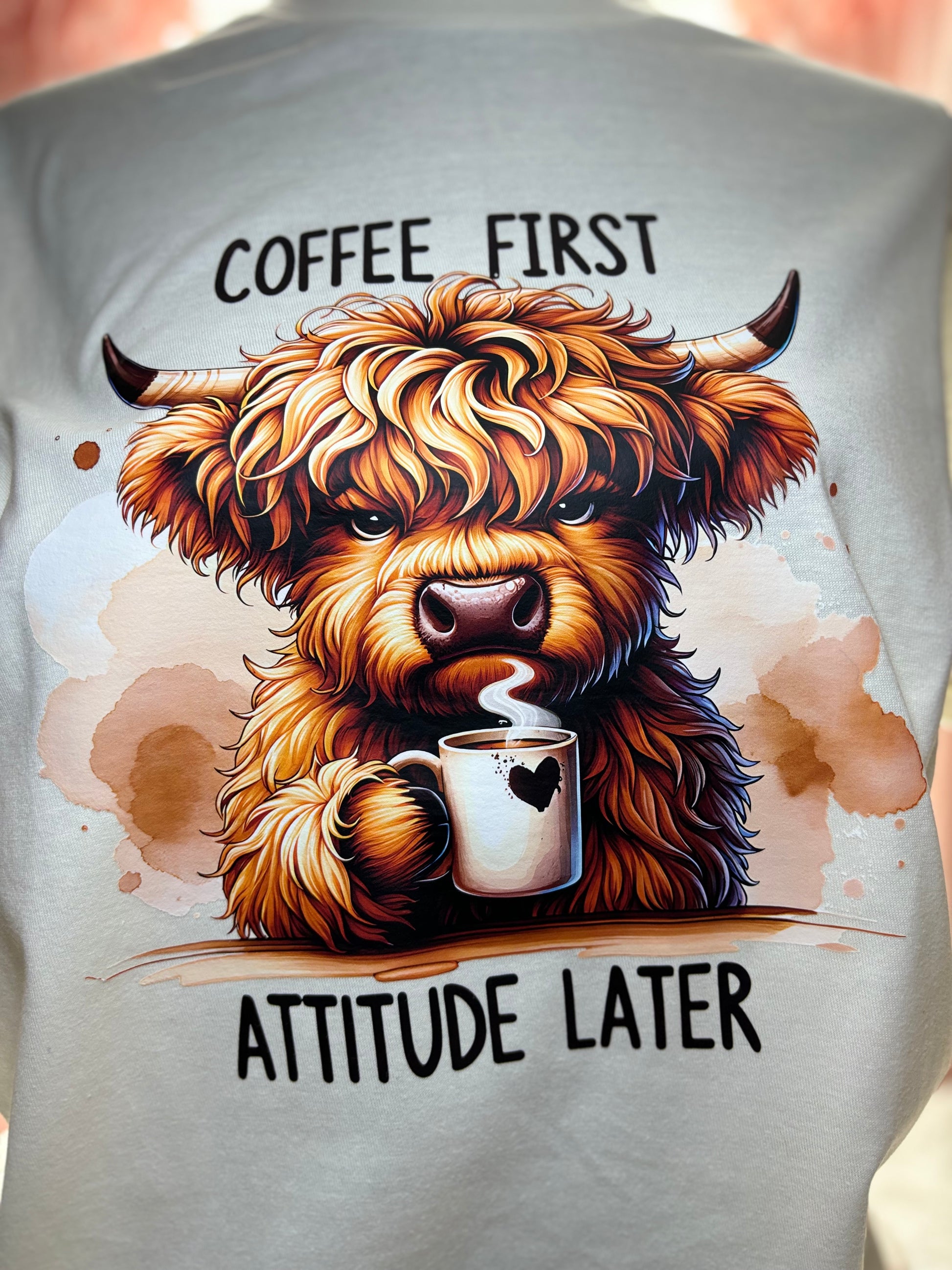Beige heat press t-shirt with a Highland cow holding a coffee cup and text 'Coffee First, Attitude Later'