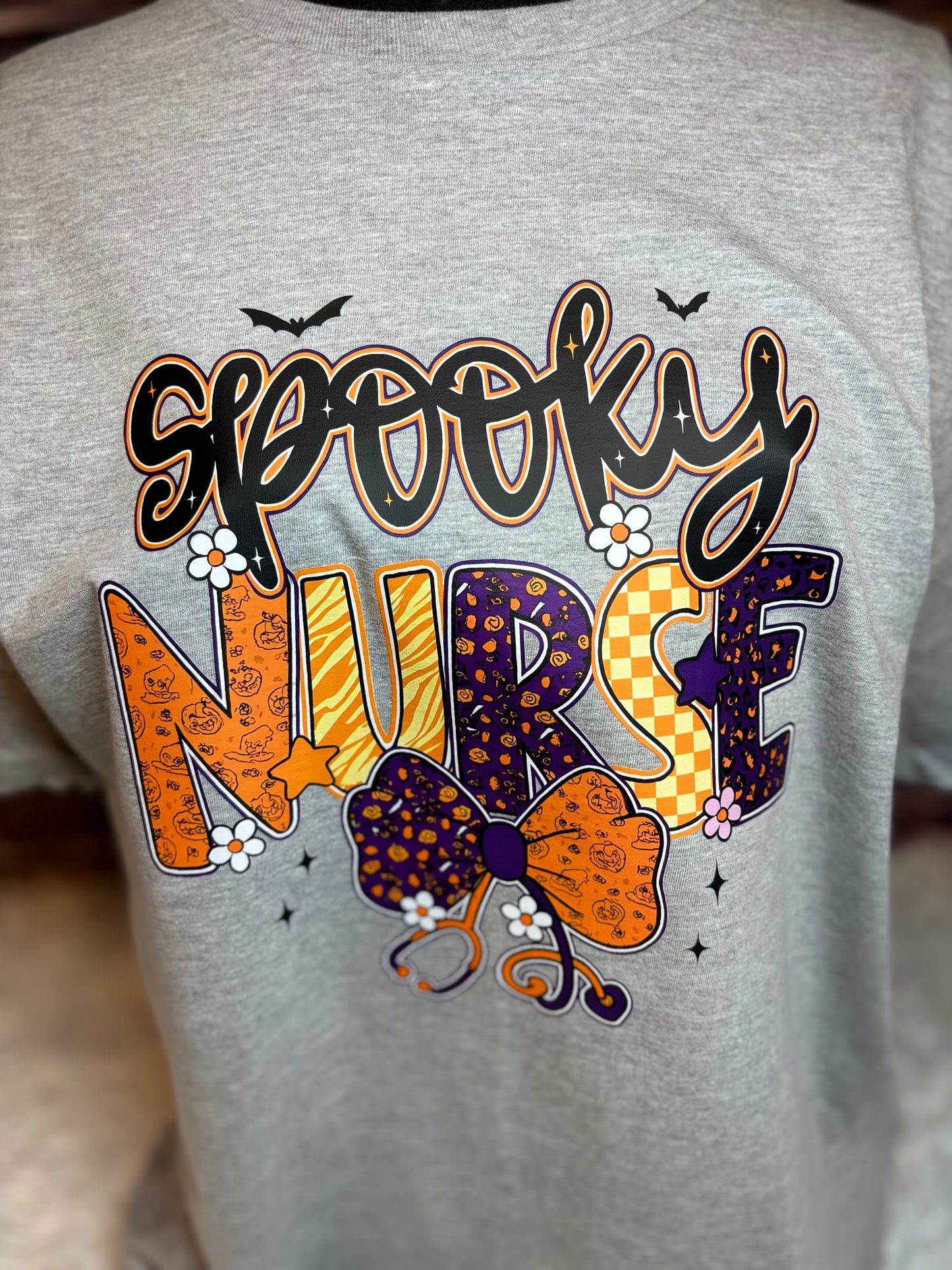 Gray heat press shirt with 'Spooky Nurse' design featuring Halloween elements- bats, stethoscope, flowers, stars