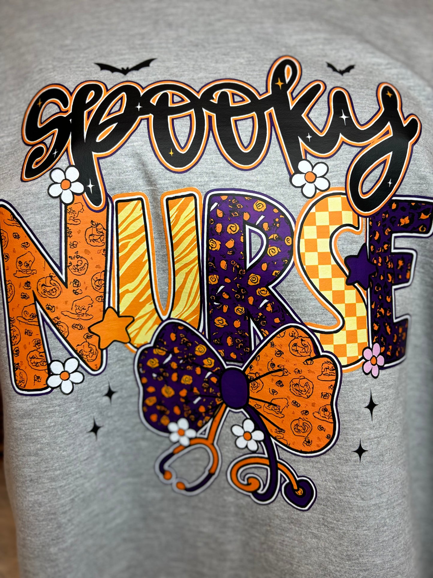 Gray heat press shirt with 'Spooky Nurse' design featuring Halloween elements- bats, stethoscope, flowers, stars