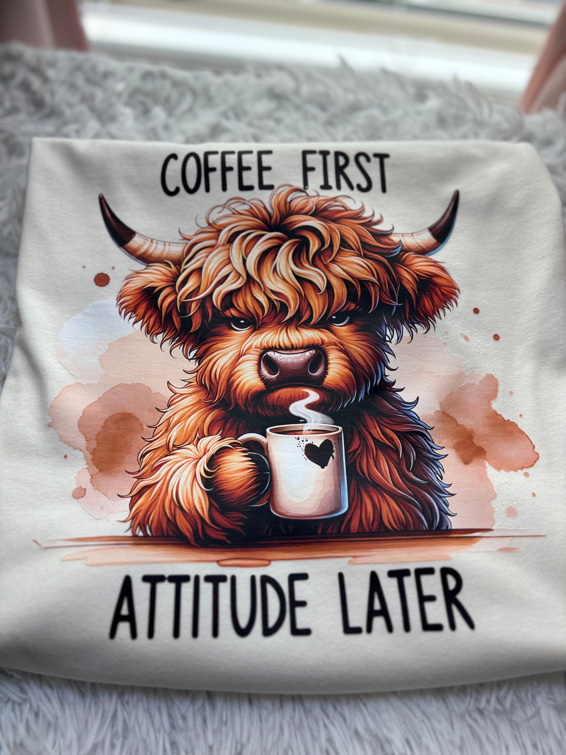 Beige heat press t-shirt with a Highland cow holding a coffee cup and text 'Coffee First, Attitude Later' 